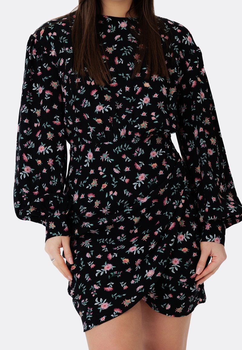 Femme portant une robe noire à manches longues avec un imprimé floral rose et vert, présentant une jupe portefeuille et des poignets froncés.