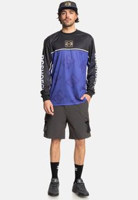 Zwarte en blauwe longsleeve trui met DC-logo, gecombineerd met grijze cargo shorts. Zwarte sokken en sneakers maken de outfit compleet.