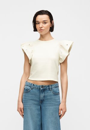 Νεαρή γυναίκα με κοντά σκούρα μαλλιά που φοράει κρεμ cropped τοπ με βολάν στα μανίκια και μπλε ψηλόμεσα τζιν, σε απλό φόντο.