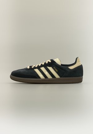 Baskets Adidas Samba noires et beiges avec trois bandes, lacets beiges et languette de talon texturée beige sur un fond neutre.