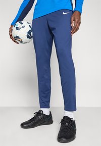 Modré športové nohavice Nike s úzkym strihom, s bielym logom, v kombinácii s čiernymi futbalovými topánkami a držiace futbalovú loptu.