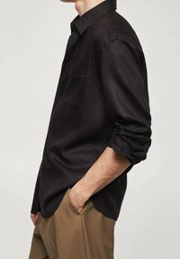 Chemise noire à manches longues en tissu lisse avec une coupe décontractée, poche poitrine unique et manches retroussées, portée avec un pantalon marron.