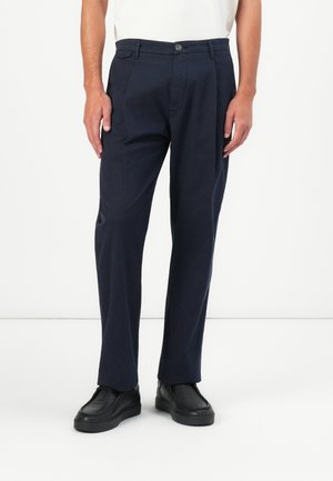 Mann trägt marineblaue gerade geschnittene Hose mit Knopf- und Reißverschluss, kombiniert mit schwarzen Slippern und einem weißen Hemd.