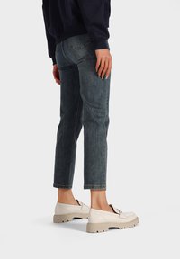 Scarpe beige slip-on con una superficie testurizzata, dotate di una suola spessa e chunky. L'outfit include jeans blu scuro e una felpa navy.