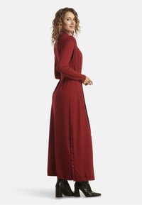 Long red dress with a high neckline, long sleeves, and side button details. Made from a soft material, paired with black heeled boots.