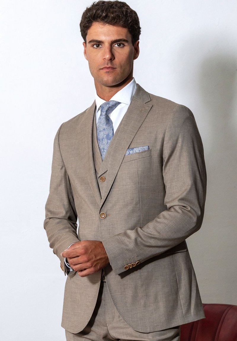 Mann trägt einen beige-einreihigen Anzug mit hellblau gemusterten Krawatte und passendem Einstecktuch, steht vor einem schlichten weißen Hintergrund.