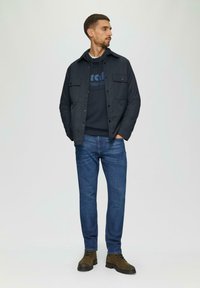 Marine knopenjack over een marine sweatshirt met blauwe tekst, gecombineerd met blauwe jeans en bruine veterschoenen. Eenvoudig en functioneel ontwerp.