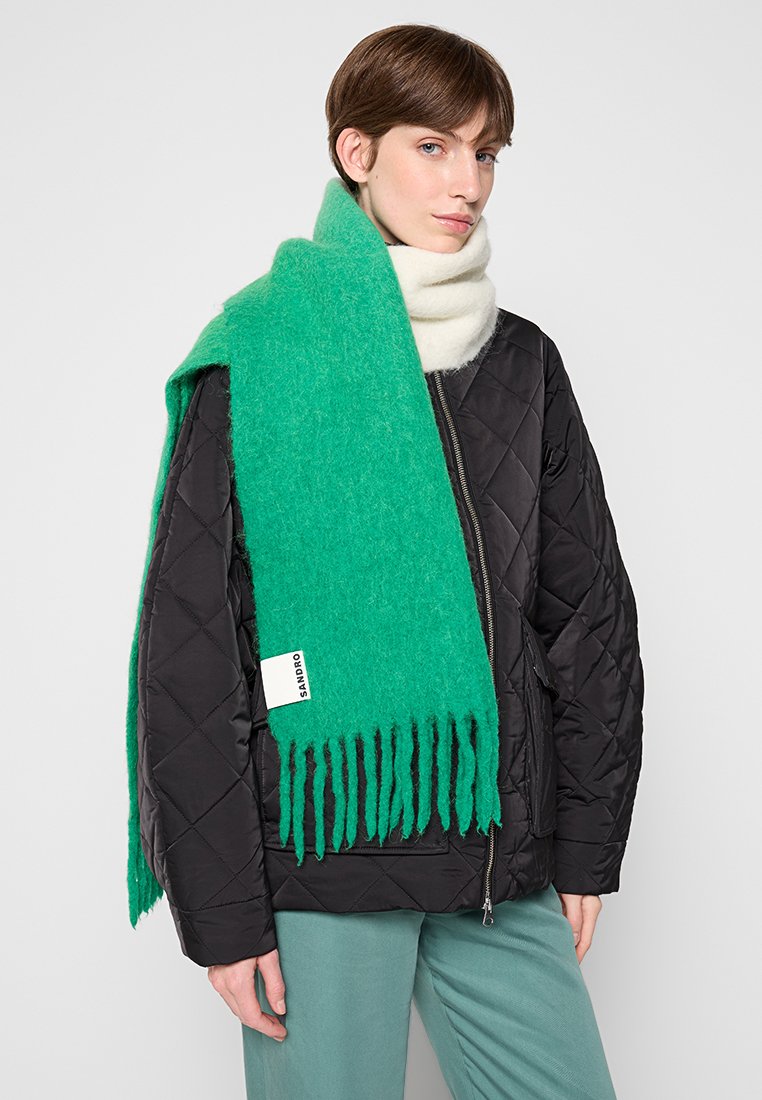 Grüner Wollschal mit Fransen, getragen über einer schwarzen gesteppte Jacke. Besitzt ein weißes Etikett mit Text. Der Stoff wirkt weich und strukturiert.