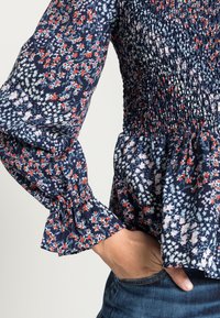 Blusa floral azul-marinho com corpinho franzido e mangas largas, apresentando flores vermelhas e rosas sobre um fundo preto. Textura do tecido macia.