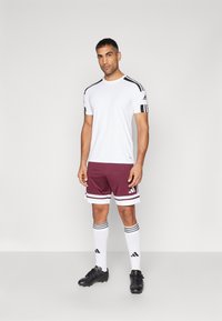Witte sportshirt met zwarte accenten, bordeauxrode shorts met witte strepen, witte sokken met zwarte strepen en zwarte voetbalschoenen.