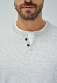 Hellgrau strukturierter Pullover mit einem Henley-Ausschnitt, der zwei braune Knöpfe aufweist. Über einem weißen T-Shirt getragen, mit dezentem Logo auf der Brust.