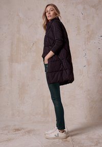 Czarna pikowana kamizelka z wysokim kołnierzem, zestawiona z zielonymi obcisłymi spodniami i białymi trampkami. Widoczne kieszenie boczne i gładka tekstura.