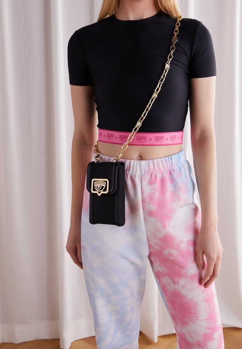 Žena v černém crop topu, růžových a modrých kalhotách s tie-dye vzorem a černé crossbody tašce se zlatým řetízkem a symbolem na sponě.