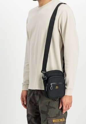 Uomo che indossa una camicia beige a maniche lunghe e pantaloni mimetici con una borsa nera a tracolla appesa alla spalla e poggiata sull'anca.