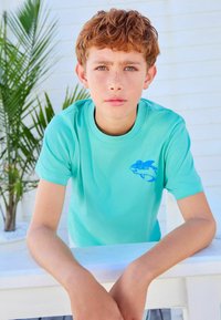 Himmelblaues T-Shirt aus Baumwolle mit rundem Halsausschnitt. Auf der linken Brust befindet sich ein grafisches Motiv eines blauen Fisches. Glatte Textur.