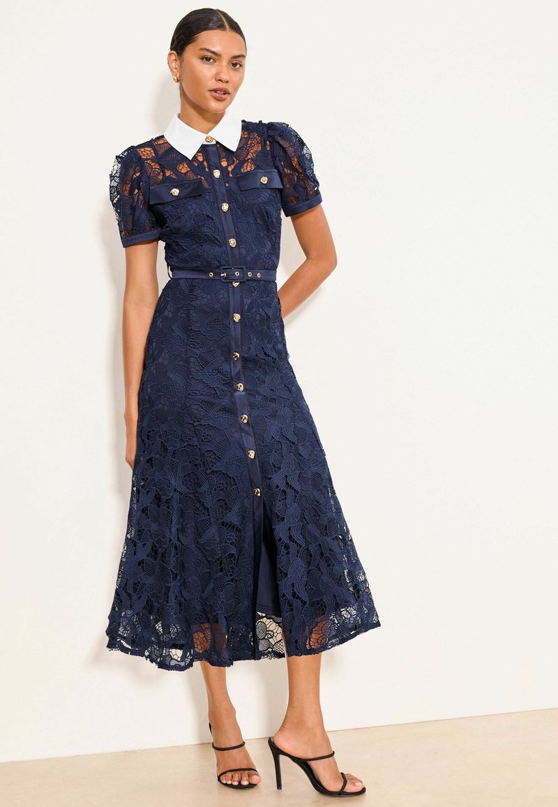 Woman wearing navy lace midi dress with white collar, short puff sleeves, front buttons, and thin belt, paired with black high-heeled sandals.