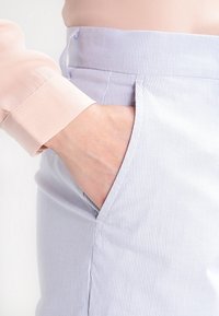 Randiga byxor med ett ljusblått och vitt mönster, med en sidoficka och ett mjukt, lätt tyg. Synlig hand i fickan.