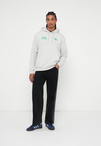 Szara bluza z kapturem z zielonym logo i emblematem "Adidas Originals", zestawiona z czarnymi spodniami i niebieskimi sneakersami z białymi paskami.