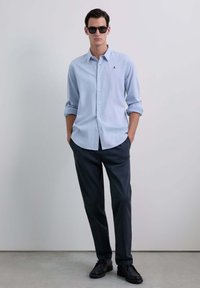 Camisa de botón azul claro con mangas largas y puños remangados, combinada con pantalones oscuros y zapatos negros; las gafas de sol que la acompañan completan el look.