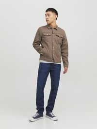 Brun denimjakke med to frontlommer og sølvknapper, parret med mørkeblå jeans og marineblå joggesko. Mot en enkel hvit bakgrunn.
