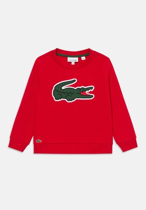 Raudona ilga rankovė megztinis su rausvais elastiniais rankogaliais, dideliu žaliu krokodilo logotipu centre priekyje ir mažesniu logotipu arčiau apačios.