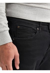 Primer plano de una persona que lleva jeans negros y una camisa de manga larga gris, mostrando la mano izquierda con cicatrices visibles cerca del área del bolsillo.