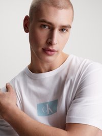 T-shirt bianca in cotone con un logo rettangolare azzurro chiaro con le lettere "cK" in bianco, con maniche corte e scollo rotondo.