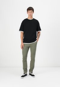 T-shirt en coton noir avec des finitions blanches, associé à un pantalon ajusté vert olive et des baskets noires avec des semelles blanches, se tenant devant un fond clair.