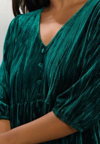 Smaragdgrünes Samtkleid mit tiefem V-Ausschnitt, kurzen puffigen Ärmeln und fünf runden Knöpfen entlang der Vorderseite, mit einer gerafften Taille.