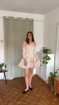 Ροζ φόρεμα με φουσκωτά μανίκια, υφασμάτινο floral σχέδιο, φουσκωτή φούστα. Μαύρες μυτερές γόβες, εσωτερικός χώρος με φυτά και ξύλινο δάπεδο.