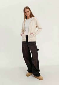 Jovem mulher com cabelo liso e longo, vestindo um casaco com fecho éclair creme, uma camisa branca, calças largas pretas e sapatos tipo slip-on bege, de pé contra um fundo liso.