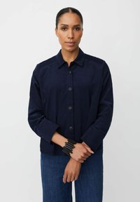 Giacca button-up blu navy in un tessuto testurizzato, con colletto e maniche lunghe. Abbinata a jeans scuri e un bracciale nero.