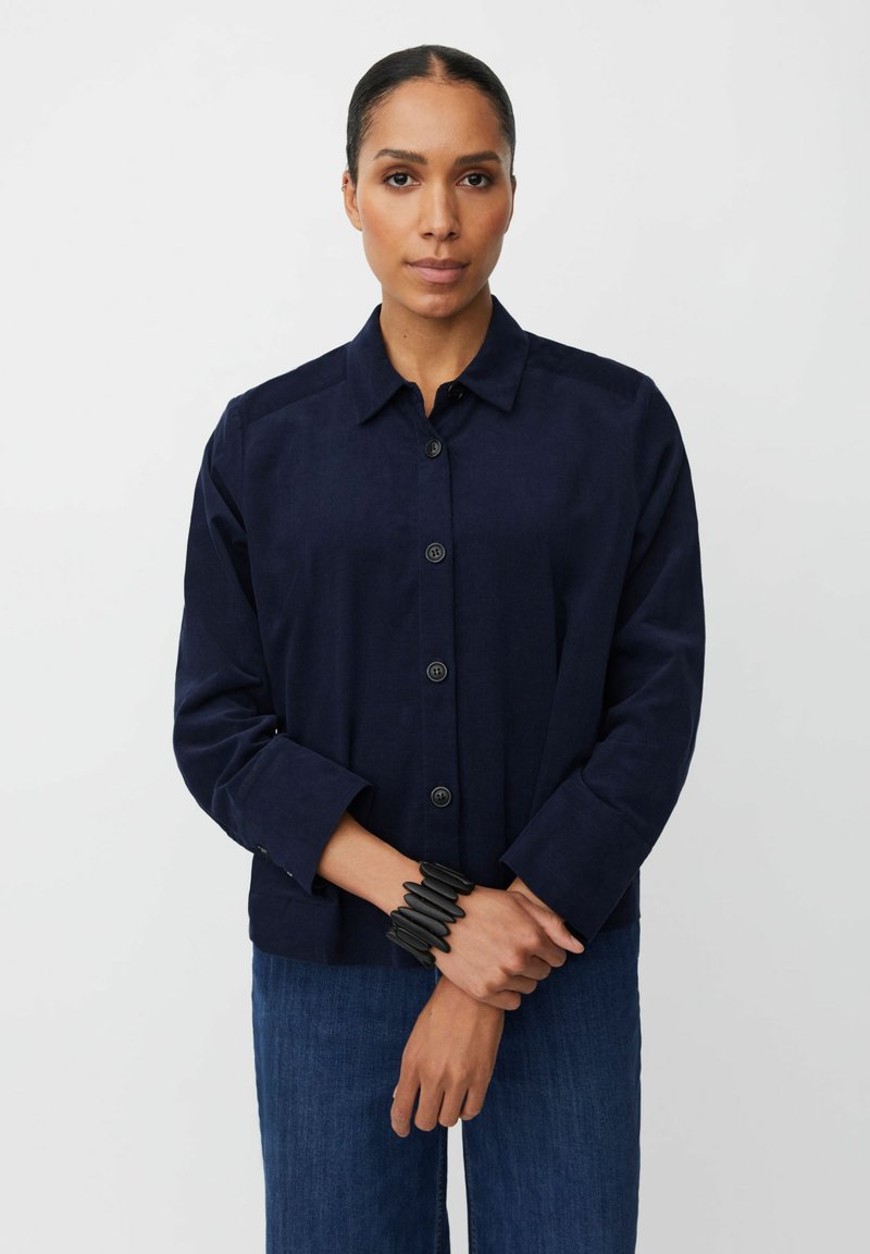 Giacca button-up blu navy in un tessuto testurizzato, con colletto e maniche lunghe. Abbinata a jeans scuri e un bracciale nero.