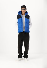 Gilet rembourré bleu avec épaules, capuche et fermeture éclair noires, porté par-dessus une chemise à manches longues blanche. Un jean noir et des baskets blanches complètent la tenue.