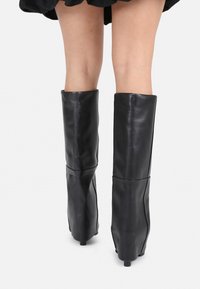 Stivali neri alti al ginocchio in pelle liscia, con un design strutturato e un dettaglio sottile di piegatura nella parte superiore, su un piccolo tacco.