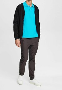 Cardigan noir porté sur un polo turquoise, associé à un pantalon foncé et des baskets blanches. Le cardigan est muni de boutons et le polo présente un col classique.