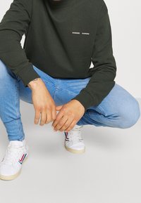 Sudadera verde oscuro con puños acanalados, con texto de logo en blanco. Jeans pitillo azul claro y zapatillas blancas con detalles en rojo y azul.