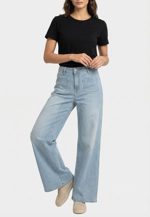 Vrouw draagt een zwart kortärmelig shirt, lichtblauwe high-waisted jeans met brede pijpen en voorzakken, en beige sneakers, staand met gekruiste benen.