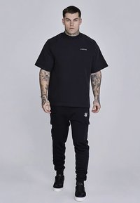 T-shirt noir oversize et joggers, en coton, avec un logo sur la poitrine et une poche. Texture lisse, coupe décontractée. Baskets noires.