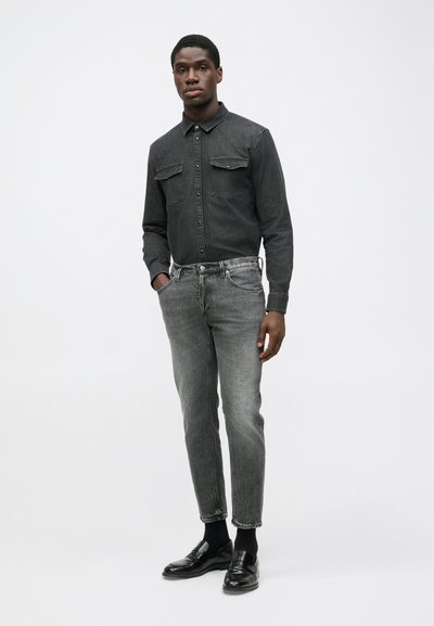 Camicia di denim scuro con design a bottoni e due tasche sul petto, abbinata a jeans grigi a gamba affusolata e mocassini neri. Stile minimalista.