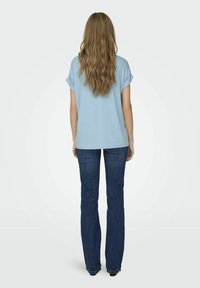 Lyseblå kortermet topp parert med mørkeblå utvidede jeans, sett fra baksiden. Toppen har en avslappet passform og jevn tekstur.