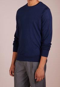 Granatowy sweter z długim rękawem, wykonany z miękkiego materiału, z bawełnianymi ściągaczami przy mankietach i u dołu. Noszony z szarymi, dopasowanymi spodniami.