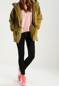 Olivgrön parka, kraftig rosa stickad tröja, svarta leggings och rosa sportskor med vita ränder. Parkan har en huva och dragsnören.