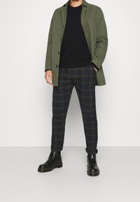 Homme portant un manteau vert olive, un pull noir, un pantalon à carreaux bleu marine et marron, et des bottes en cuir noir, debout les mains dans les poches.