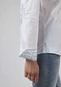 Chemise blanche avec des accents floraux sur les manches et l'ourlet, présentation d'un tissu texturé et d'un patch arrondi au niveau du coude.