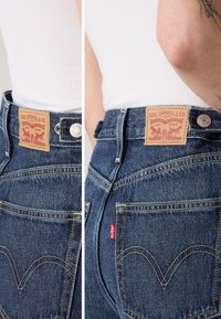 Jean en denim bleu foncé avec un patch en cuir marron portant un graphique rouge. Ils sont dotés de coutures jaunes, de matériel en métal argenté et de poches arrière bien visibles.