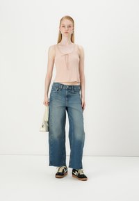 Light pink ribbed tank top with a tie detail, paired with wide-leg faded blue jeans. Model holds a small white handbag, wearing black sneakers.