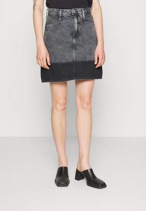 Žena v dvoubarevné mini sukni z černého a šedého denimu, v černém tílku bez rukávů a v černých lodičkách bez zadního pásku, stojící na světlé podlaze.