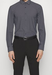 Getailleerd herenshirt in donker marineblauw met een diamantpatroon, lange mouwen en een button-down kraag. Gecombineerd met een zwarte broek en een riem.