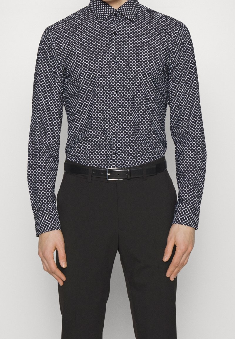 Camisa ajustada de hombre en azul marino oscuro con un patrón de diamantes, mangas largas y cuello abotonado. Combinada con pantalones negros y un cinturón.